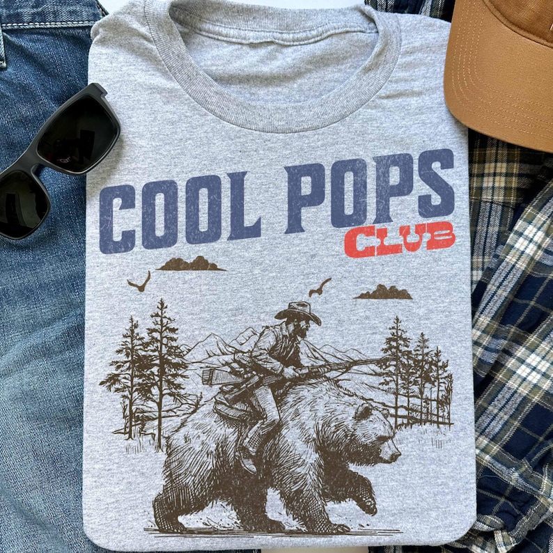 May include: Gray t-shirt with a vintage-style graphic of a cowboy riding a bear. The text "COOL POPS CLUB" is printed in blue and red on the shirt.