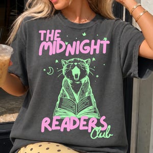 May include: A charcoal gray short-sleeve t-shirt featuring a graphic of a yawning cat reading an open book. The text reads THE MIDNIGHT READERS CLUB in pink and light green. Small stars and a crescent moon surround the cat in light green ink.
