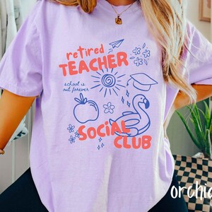 May include: A light purple t-shirt with the words "retired TEACHER SOCIAL CLUB" in red and blue. The shirt has a graphic of a sun, apple, flamingo, and other small illustrations. The word "orchid" is written in white at the bottom right.