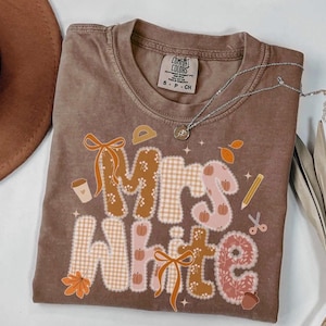 May include: A brown short-sleeve t-shirt featuring the text Mrs. White in large, patterned letters. The design includes graphics of pumpkins, leaves, a pencil, scissors, a coffee cup, and bows. The shirt has a Comfort Colors brand label.