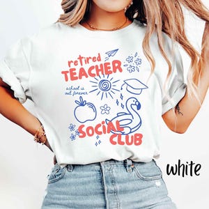 May include: White t-shirt with a graphic design in red and blue. The design includes the words "retired TEACHER SOCIAL CLUB" with illustrations of a sun, apple, graduation cap, and a flamingo floatie. The text "school is out forever" is also included.