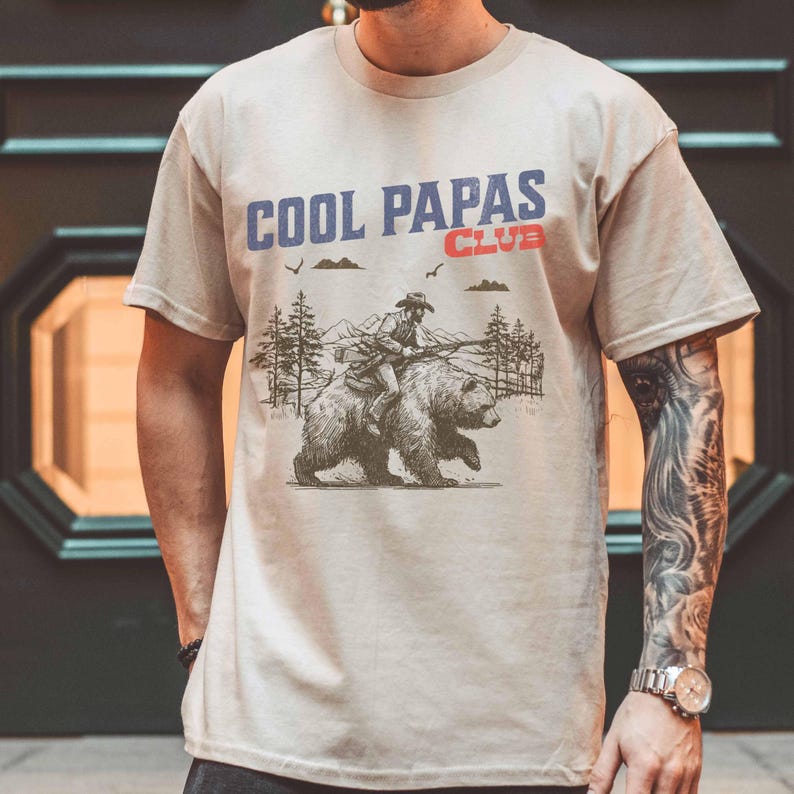 May include: A beige t-shirt featuring a graphic print of a person riding a bear. The text "COOL PAPAS CLUB" is printed in blue and red on the shirt.