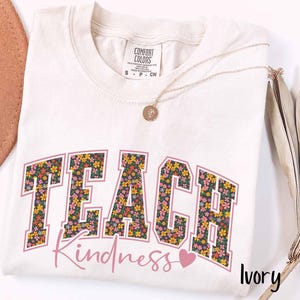 May include: An ivory-coloured t-shirt with the words "TEACH Kindness" in a floral design. The word "TEACH" is in a block letter style, and "Kindness" is in a cursive font. A gold necklace is draped over the shirt.