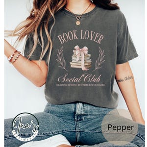 May include: A gray t-shirt with the text "Book Lover Social Club" and a stack of books tied with a pink ribbon. The text "Reading beyond bedtime encouraged" is below the graphic.