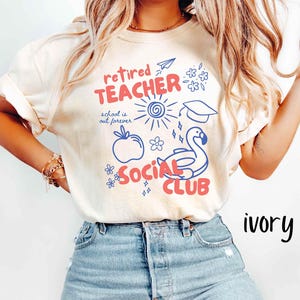 May include: Ivory-colored t-shirt with a graphic design in red and blue. The design includes the words "Retired Teacher Social Club" with illustrations of a sun, apple, graduation cap, and a swan.