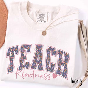 May include: Ivory-coloured t-shirt featuring the word "TEACH" in a floral design, and the word "Kindness" in a cursive font. A small pink heart is next to the word "Kindness". A gold necklace and a brown hat are also in the image.
