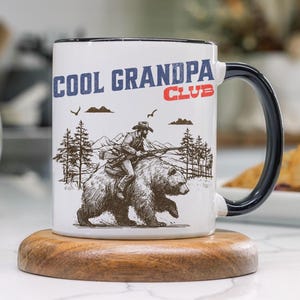 May include: White ceramic mug with a black handle and rim. The mug features the text "COOL GRANDPA CLUB" in blue and red, along with a graphic of a man riding a bear with a rifle. The mug sits on a wooden coaster.