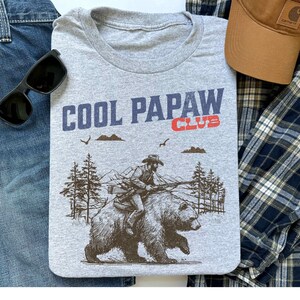 May include: Gray t-shirt with the text "COOL PAPAW CLUB" in navy and red. The graphic depicts a man riding a bear through a forest. Sunglasses, a brown cap, and a plaid shirt are also visible.