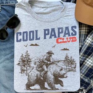 May include: Gray t-shirt with a graphic of a cowboy riding a bear. The text "COOL PAPAS CLUB" is printed on the shirt in blue and red.