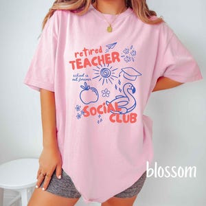 May include: A light pink t-shirt with a graphic design that reads "retired TEACHER SOCIAL CLUB". The design includes a sun, apple, flamingo, and other illustrations in blue and red. The text "school is out forever" is also included.