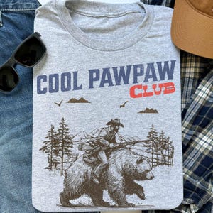 May include: Gray t-shirt with the text "Cool Pawpaw Club" in blue and red. A cartoon illustration of a man riding a bear through a forest is below the text.