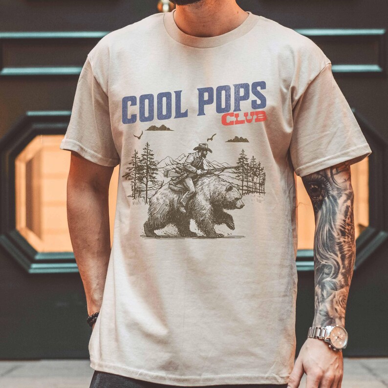May include: A beige t-shirt with a graphic print of a cartoon cowboy riding a bear. The text "COOL POPS CLUB" is printed in red and blue letters on the shirt.