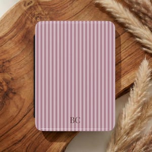 May include: A rectangular e-reader case featuring a vertical striped pattern in shades of dusty rose and light pink. The center bottom of the case displays the monogram letters BC in a simple, dark serif font. The case has rounded corners.