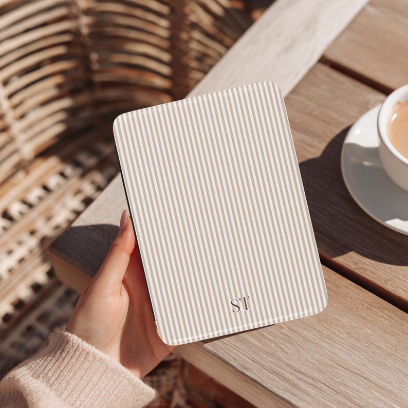 May include: A rectangular, beige and white striped phone case with the initials "ST" in the bottom right corner. The case is held by a person, with a wooden table and a cup of coffee in the background.