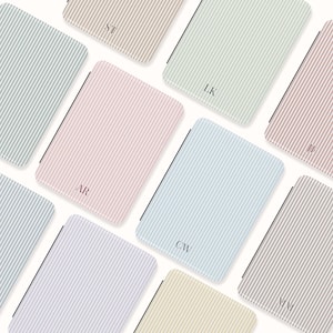 May include: An assortment of rectangular notebooks with vertical striped patterns in various pastel colors, including pink, blue, and beige. Each notebook features a different set of initials in a matching color. The notebooks are arranged on a white background.