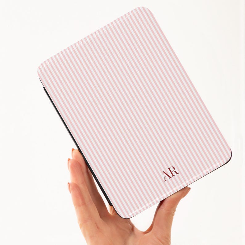 May include: A rectangular case with a pink and white striped pattern. The case has a black border and the initials "AR" in a burgundy color. The case is held in a person's hand.