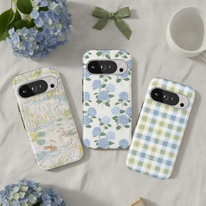 Può includere: Tre custodie per telefono con design unici. Una custodia presenta una scena di stagno con anatre, un'altra ha fiori di ortensia blu su sfondo bianco e la terza ha un motivo a quadri blu e verdi. Le custodie sono disposte su una superficie bianca.