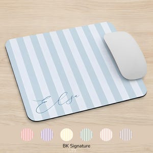 Puede incluir: Una alfombrilla de ratón rectangular con un patrón de rayas verticales en azul claro y blanco, con el nombre Elsa escrito en letra cursiva azul oscuro. Un ratón de ordenador blanco descansa sobre el lado derecho de la alfombrilla redondeada.