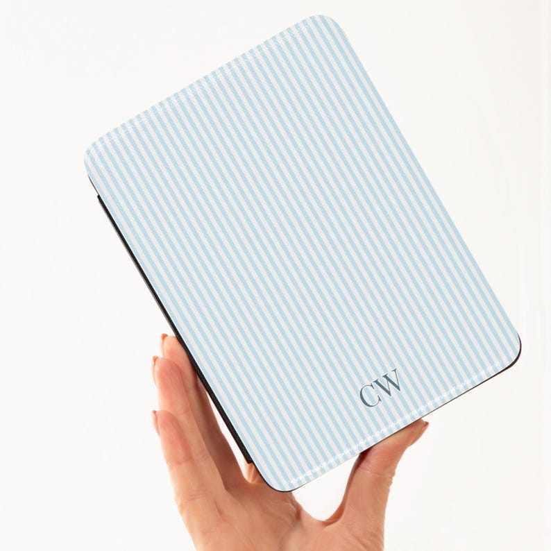 May include: A light blue and white striped tablet case with the initials "CW" in the lower right corner. The case is held in a person's hand against a white background. The case has a black border.