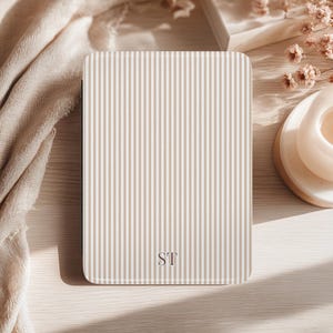 May include: A rectangular, beige and white striped case with the initials "ST" at the bottom. The case is on a light wooden surface, with a beige cloth and decorative elements in the background.