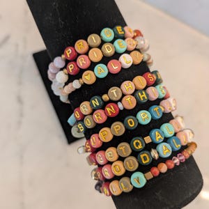 May include: A collection of colorful beaded bracelets with gold lettering spelling out phrases like "PRIDE," "ALLY," and "SUPPORT ALL." The bracelets are displayed on a black stand, showcasing a variety of colors and bead sizes.