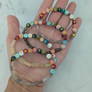 May include: Four beaded bracelets spelling out "EQUALITY FOR ALL" in colorful letter beads. The bracelets are in various colors, including red, blue, and yellow, with gold-colored accents. The bracelets are held in a hand.