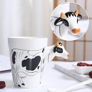 May include: A white ceramic mug with a black and white cow design. The mug has a 3D cow handle.