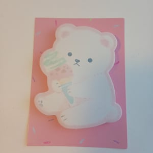 以下が含まれることがあります： アイスクリームを持った白いクマのイラストが描かれたピンク色の付箋。クマは青い目とピンクの耳をしています。背景はピンク色で、カラフルなスプリンクルが散りばめられています。