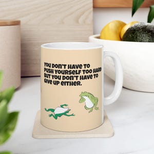以下が含まれることがあります： ベージュの背景に「YOU DON'T HAVE TO PUSH YOURSELF TOO HARD BUT YOU DON'T HAVE TO GIVE UP EITHER」というテキストがプリントされた白いセラミックマグ。マグには2匹の緑色のカエルのイラストも描かれています。マグには白いハンドルが付いています。