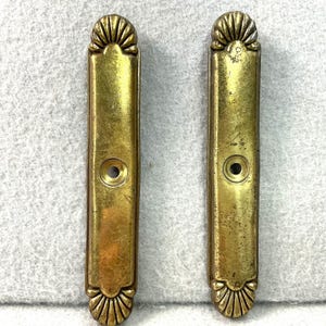 May include: Two vintage brass drawer pulls. Each pull is rectangular with a decorative fan-shaped design at the top and bottom. A single hole is centered on each pull. The brass has a warm, aged patina.