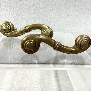 May include: Two antique brass drawer pulls with a scroll design. The pulls have a tarnished, aged appearance, with a circular end and a curved handle. The hardware is likely for a cabinet or drawer.