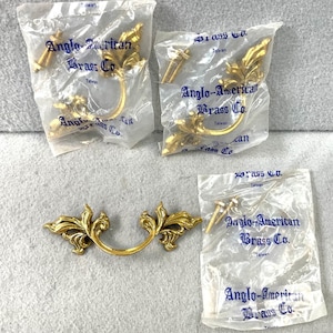 May include: Several gold-colored drawer pulls with ornate leaf designs are displayed. Some are packaged in clear plastic bags with the text "Anglo-American Brass Co. Taiwan". The pulls have a curved handle and decorative ends.
