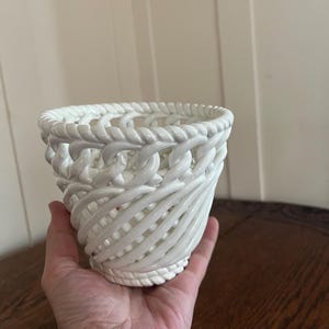 May include: A white ceramic basket-style vase with a woven rope design. The vase has a cylindrical shape with a flared top and a textured surface. The basket is held in a hand.
