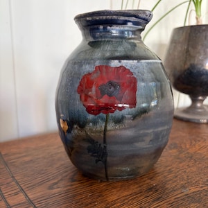 May include: A rounded ceramic vase with a glossy finish, featuring a dark blue and grey mottled glaze. A single vibrant red poppy flower with a dark center and a thin green stem is painted on the front surface of the vessel.