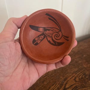 May include: A small, round, terracotta-colored clay bowl featuring a black painted graphic design in the center. The design consists of a stylized, abstract shape with a spiral element and geometric lines, set against the matte reddish-brown surface.
