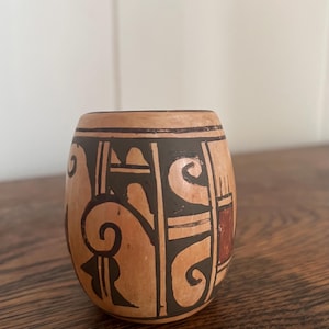 May include: A small, oval-shaped ceramic vessel with a light brown base and dark brown and red geometric patterns. The design includes swirls, lines, and rectangular shapes. The vessel is likely handmade and has a rustic aesthetic.