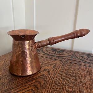 May include: A hammered copper coffee pot with a wide, flared rim and a pouring spout. The pot features a long, turned wooden handle attached with a metal bracket. The exterior surface has a distinct dimpled texture and a warm, metallic finish.