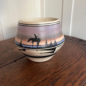 May include: A ceramic bowl with a Native American design. The bowl features a cream-colored rim and a painted scene of a rider on a horse, with a landscape in black, blue, and shades of purple and brown. The bowl has a black and white geometric pattern.