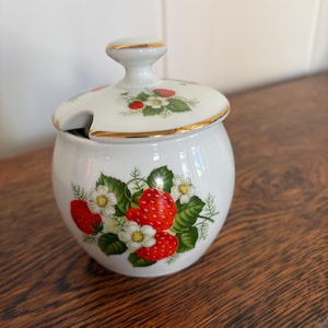 May include: A white porcelain sugar bowl with a matching lid featuring a gold-trimmed rim and a cutout for a spoon. The body and lid are decorated with a colorful botanical pattern of red strawberries, green leaves, and small white flowers.