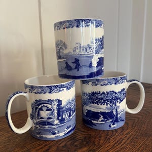 May include: Three white ceramic mugs featuring a blue transferware pattern. Each mug displays a different detailed landscape scene with figures, trees, and architectural elements, finished with a decorative floral border around the top rim.
