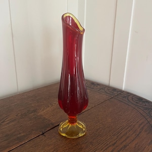 May include: A tall, red glass vase with a flared opening and a yellow rim. The vase has a bulbous base and a yellow, scalloped foot. The vase is approximately 10 inches tall.