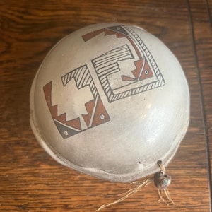 May include: A round, off-white ceramic bowl with geometric designs in brown and black. The bowl has a small hole with a string and bead attached. The bowl is sitting on a wooden surface.
