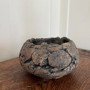 May include: A small, round, brown ceramic bowl with a textured, organic surface. The bowl has a dark interior and a rough, uneven exterior, suggesting a handcrafted or natural aesthetic. The bowl is sitting on a wooden surface.