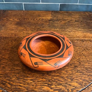 May include: A round, terracotta-colored pottery bowl with a central circular opening. The bowl features a black geometric design around the rim and exterior. The bowl is displayed on a wooden surface.
