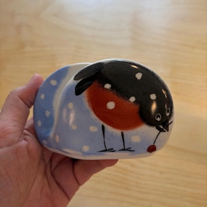 May include: A ceramic napkin holder featuring a painted bullfinch bird with a black head, orange-red chest, and white spots on a light blue background. The bird is depicted standing in snow, holding a small red berry in its beak.