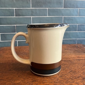May include: Cream-colored ceramic pitcher with a black rim and handle. The pitcher features a brown and black band around the base. The pitcher is on a wooden surface.