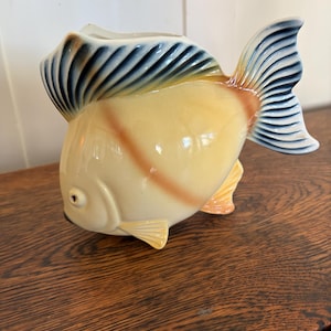 May include: A ceramic fish-shaped pitcher featuring a cream-colored body with a subtle orange vertical stripe. The dorsal and caudal fins are accented with a gradient of blue and white. The piece has a wide, open mouth at the top for pouring.