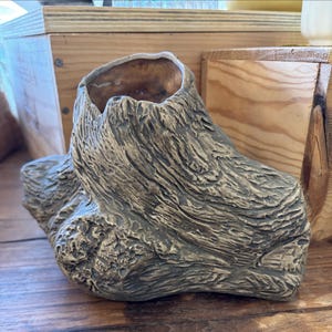 May include: A ceramic planter shaped like a weathered tree stump with a textured, bark-like surface in shades of brown and gray. The vessel features a hollow, rounded opening at the top with a smooth, light brown interior finish.