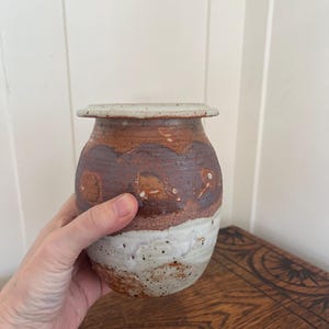 May include: A handmade ceramic vase with a wide, rounded body and a small, flat rim. The vase features a speckled white base, a band of dark brown, and a lighter brown top. The vase is held by a hand.