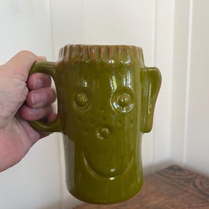 May include: A glossy olive green ceramic mug featuring a stylized face with raised circular eyes, a small nose, and a curved mouth. The mug has a textured rim, a single handle on the left side, and a protruding ear-like shape on the right side.